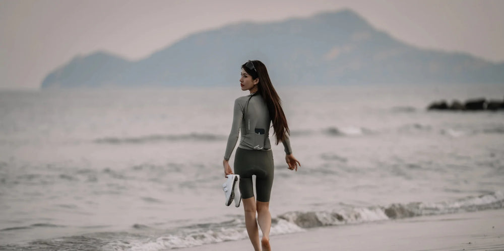 A woman in a gray long-sleeved wetsuit walks barefoot holding swim fins for the 20th anniversary special bib shorts article
