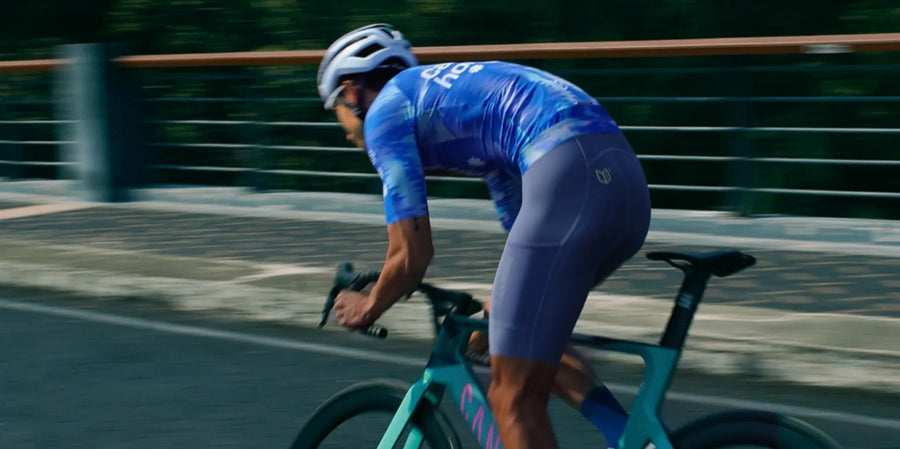 Ein Radfahrer trägt ein blau-weißes Trikot in Kombination mit lila Radshorts und fährt ein blaugrünes Straßenrad aus der Neuheitenkollektion für Herren.