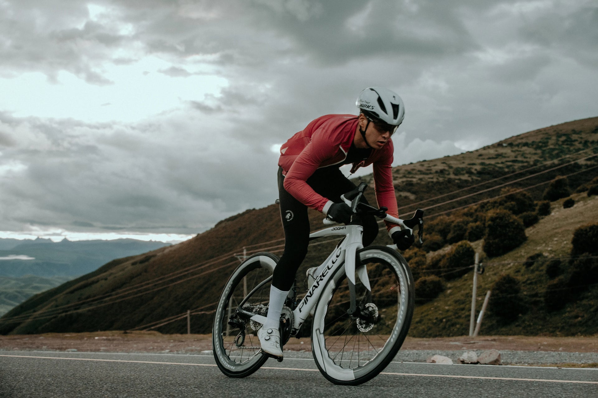Cyclist in a red jacket riding on a mountain road with a cloudy sky.