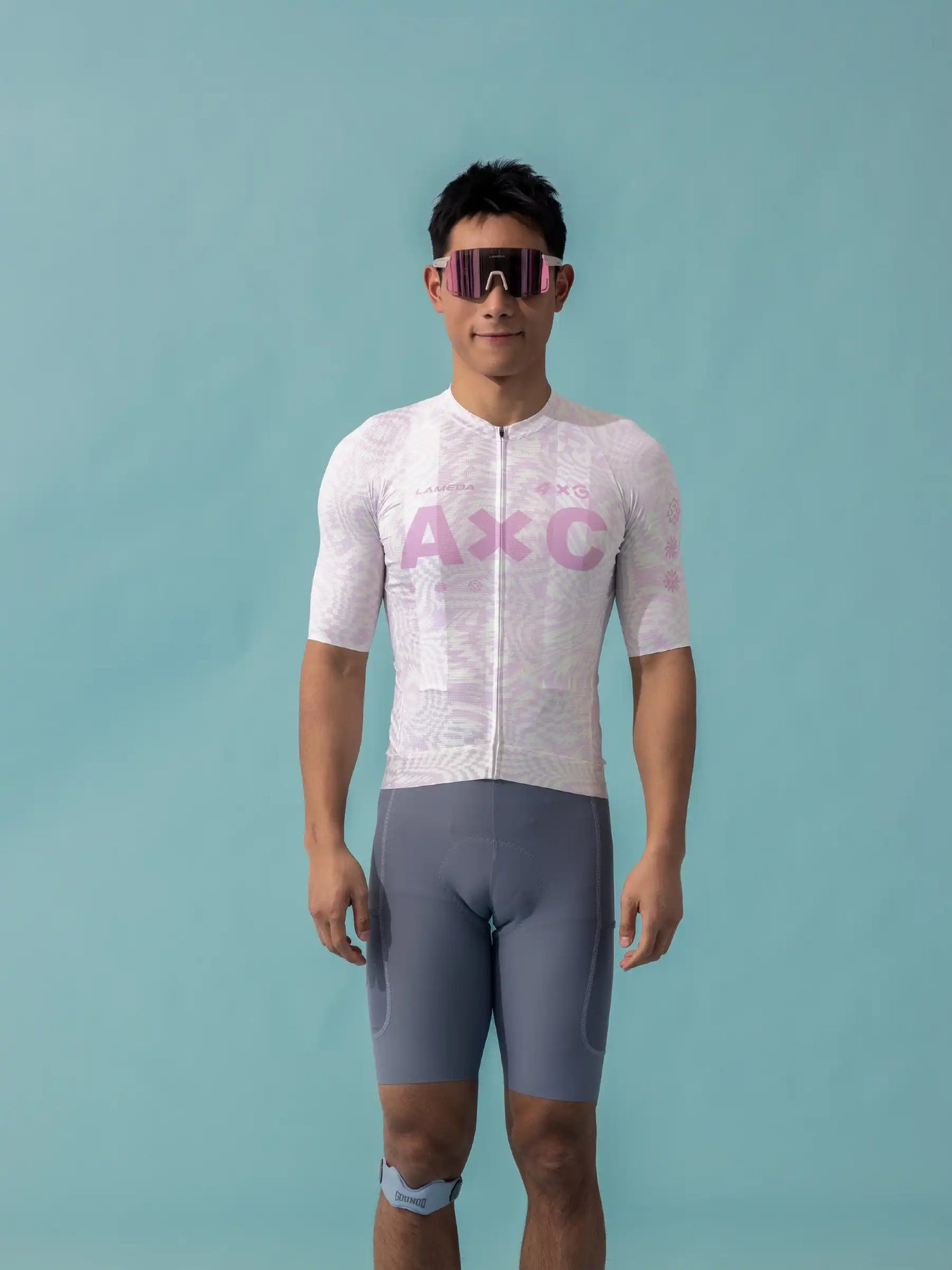 En man bär en vit- och rosa mönstrad LAMEDA x australian studio unisex cykeltröja med grå kompressionsshorts